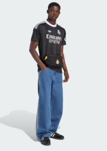 Young man in a black Real Madrid jersey with a waist bag across his hips, wearing loose blue jeans and black loafers, standing against a light background.