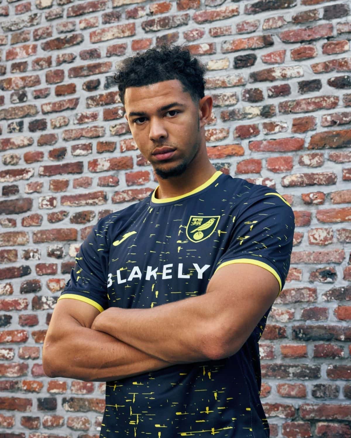 Young man with short curly hair standing with arms crossed in a navy yellow soccer jersey against a red brick wall in the background.