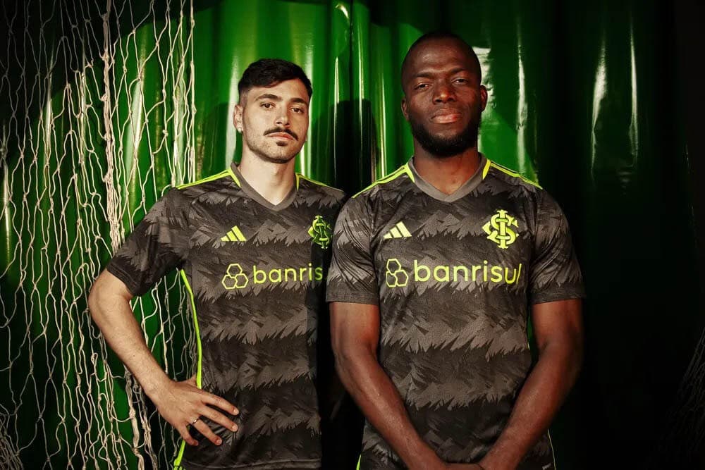 1MQbkVfEDXNCy1c_bfb8cf8c-7e95-43be-a8f7-c5ded19747ea | UAE SPORT STORE Two male soccer players in matching dark camouflage jerseys with neon green accents stand side by side against a green backdrop, posing for a photo.
