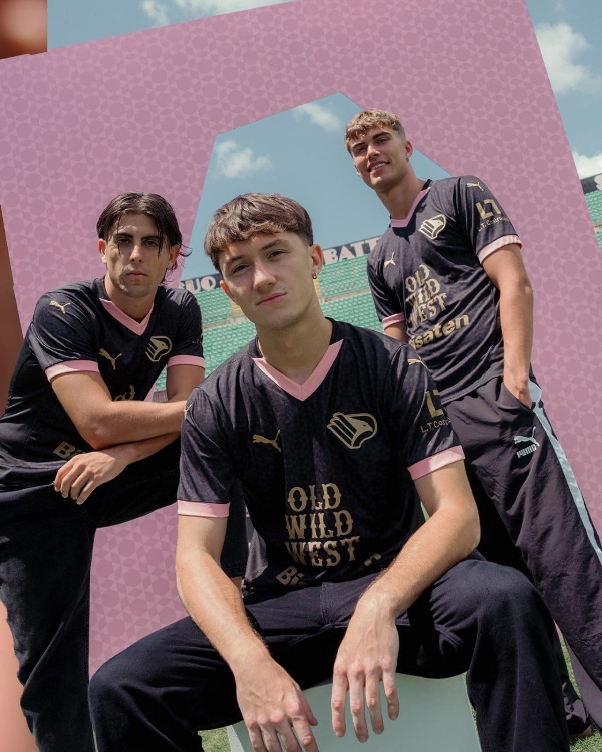 Three young men in black soccer jerseys with pink trim posing together on a field, framed by a pink geometric structure.