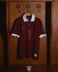 Maroon polo shirt with white collar hanging on a wooden wardrobe, Torino FC emblem visible at the bottom.