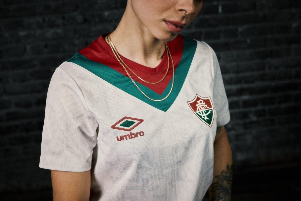 Close-up of a person wearing a white sports jersey with a green and red V-neck, showing a crest and Umbro logo, plus gold chains and a nose ring against a dark brick wall background.