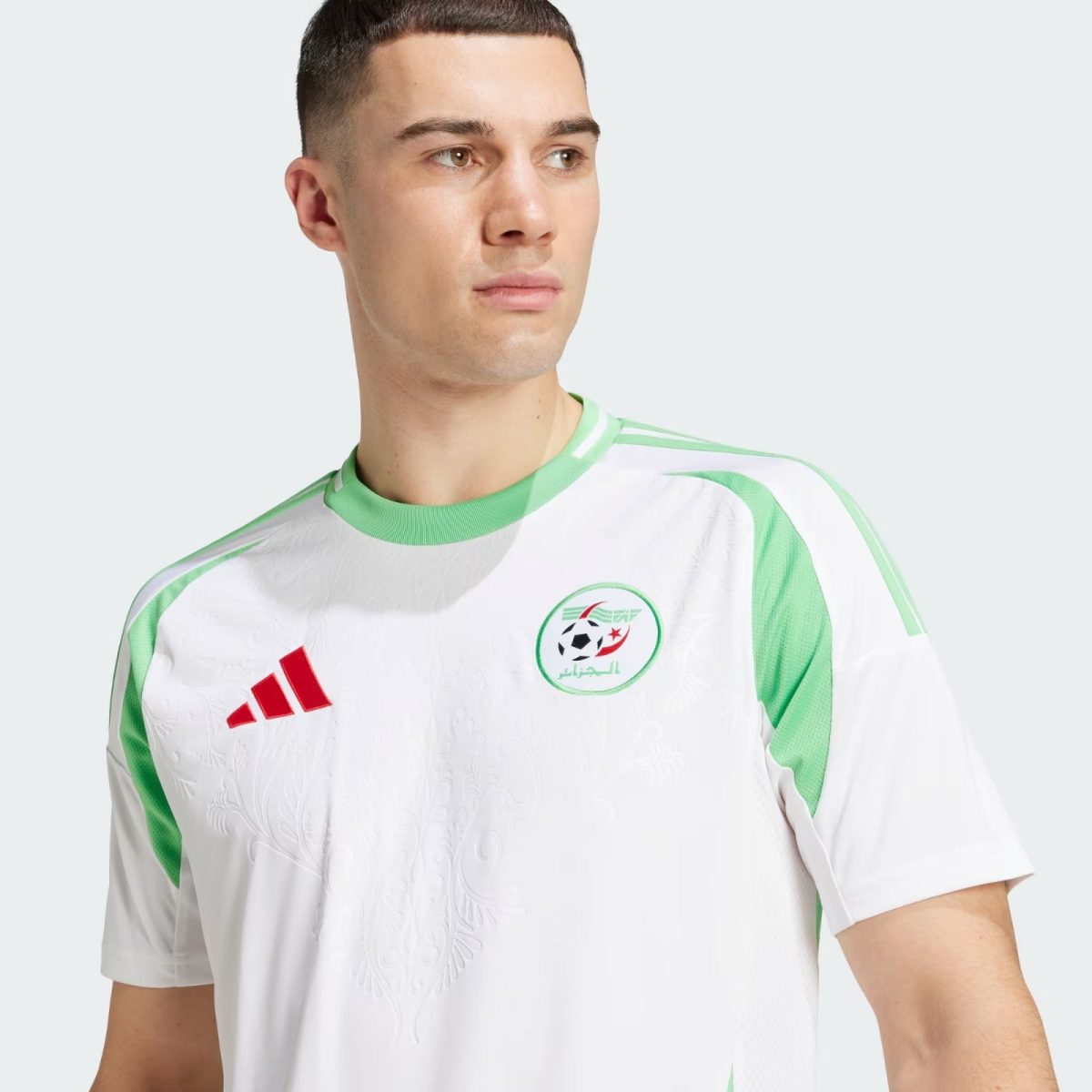 Young man wearing a white and green Algeria national team jersey, looking to the right, against a pale background.