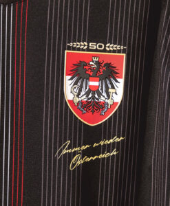 A badge showing the Austrian black eagle on a red‑and‑white shield with a crown, with a gold '50' above, mounted on a dark pinstripe suit and gold cursive text beneath.