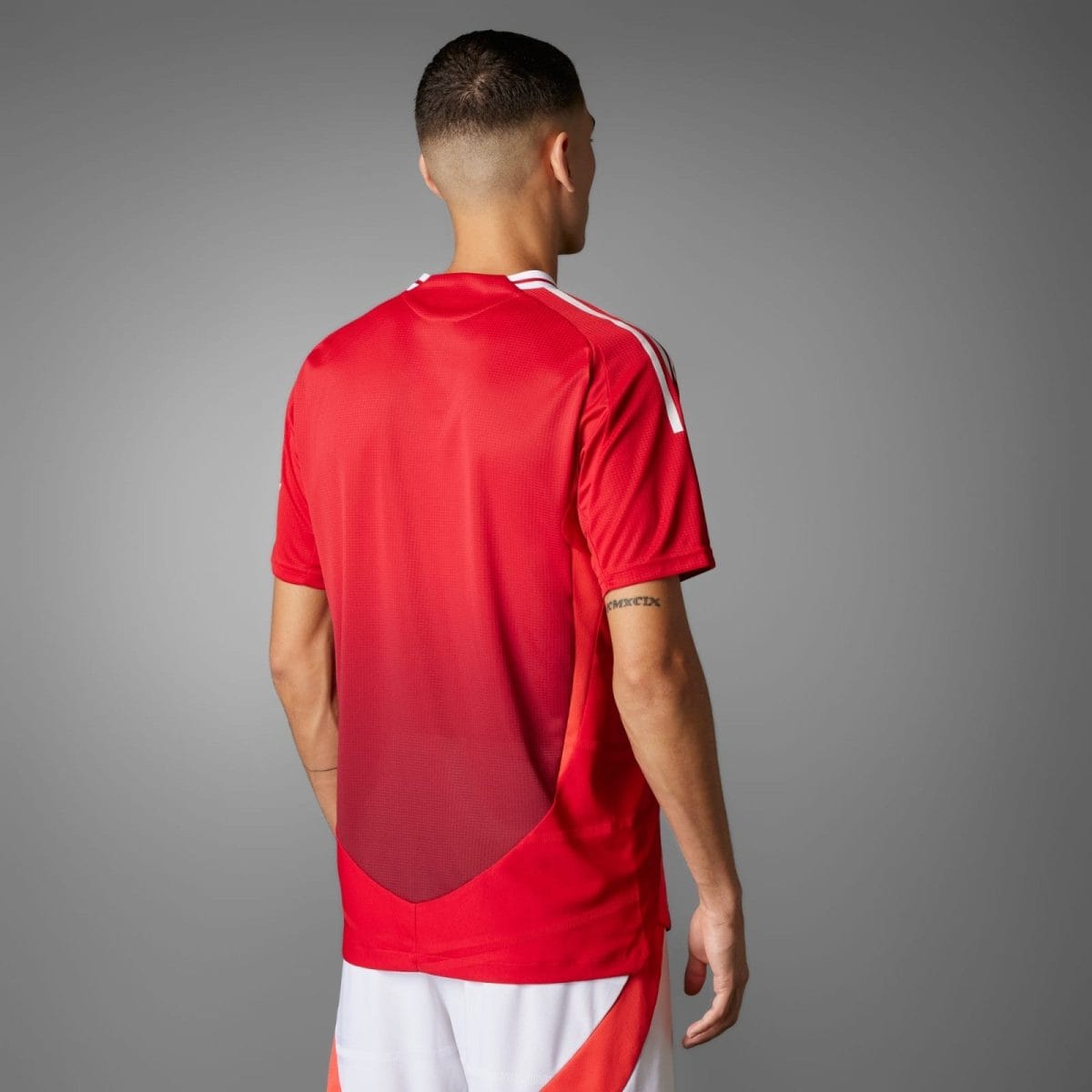 Man in a red athletic jersey seen from behind, fade haircut, standing against a gray studio backdrop.