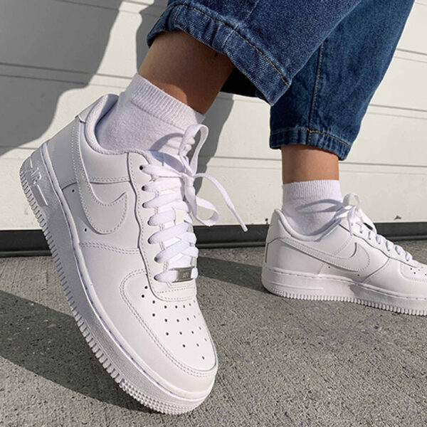 Pair of white sneakers being worn with rolled jean cuffs and white socks, standing on concrete pavement in sunlight.