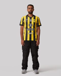 Front-facing man wearing a yellow and black striped soccer jersey with logos against a gray background.