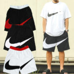 Man wearing a white Nike T-shirt standing beside black and white Nike shorts with oversized swoosh logos; multiple shorts are displayed nearby.