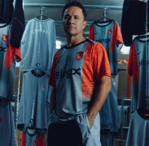 Man in a red-and-gray sports jersey stands in a locker room surrounded by hanging jerseys and equipment in the background.