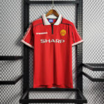 Red Manchester United home jersey hanging on a wooden hanger against a metal rack, featuring SHARP sponsor and Umbro logo with club crest.