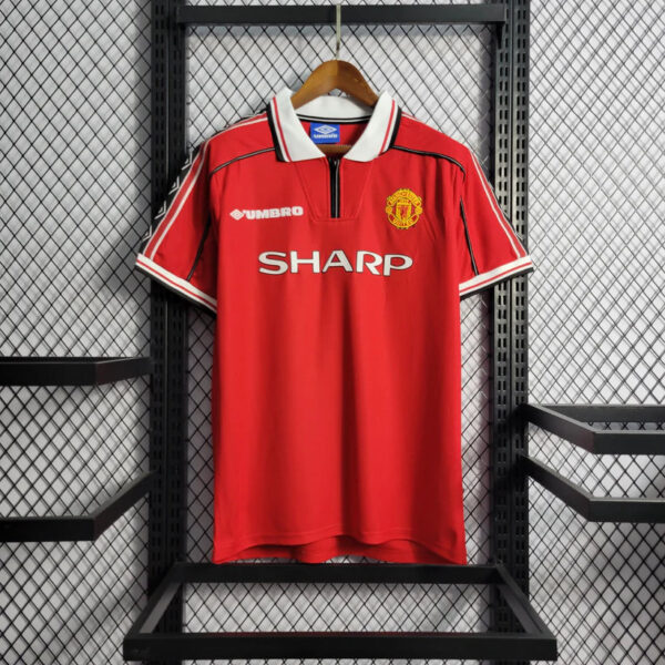 Red Manchester United home jersey hanging on a wooden hanger against a metal rack, featuring SHARP sponsor and Umbro logo with club crest.