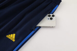 1a6ac6ad | UAE SPORT STORE Navy Adidas track pants with a yellow logo and blue zipper, a smartphone peeking out of the side pocket in a flat lay.