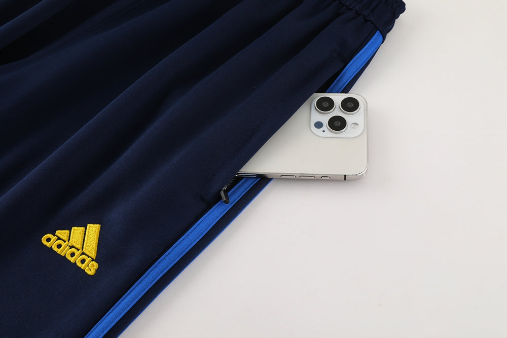 1a6ac6ad | UAE SPORT STORE Navy Adidas track pants with a yellow logo and blue zipper, a smartphone peeking out of the side pocket in a flat lay.