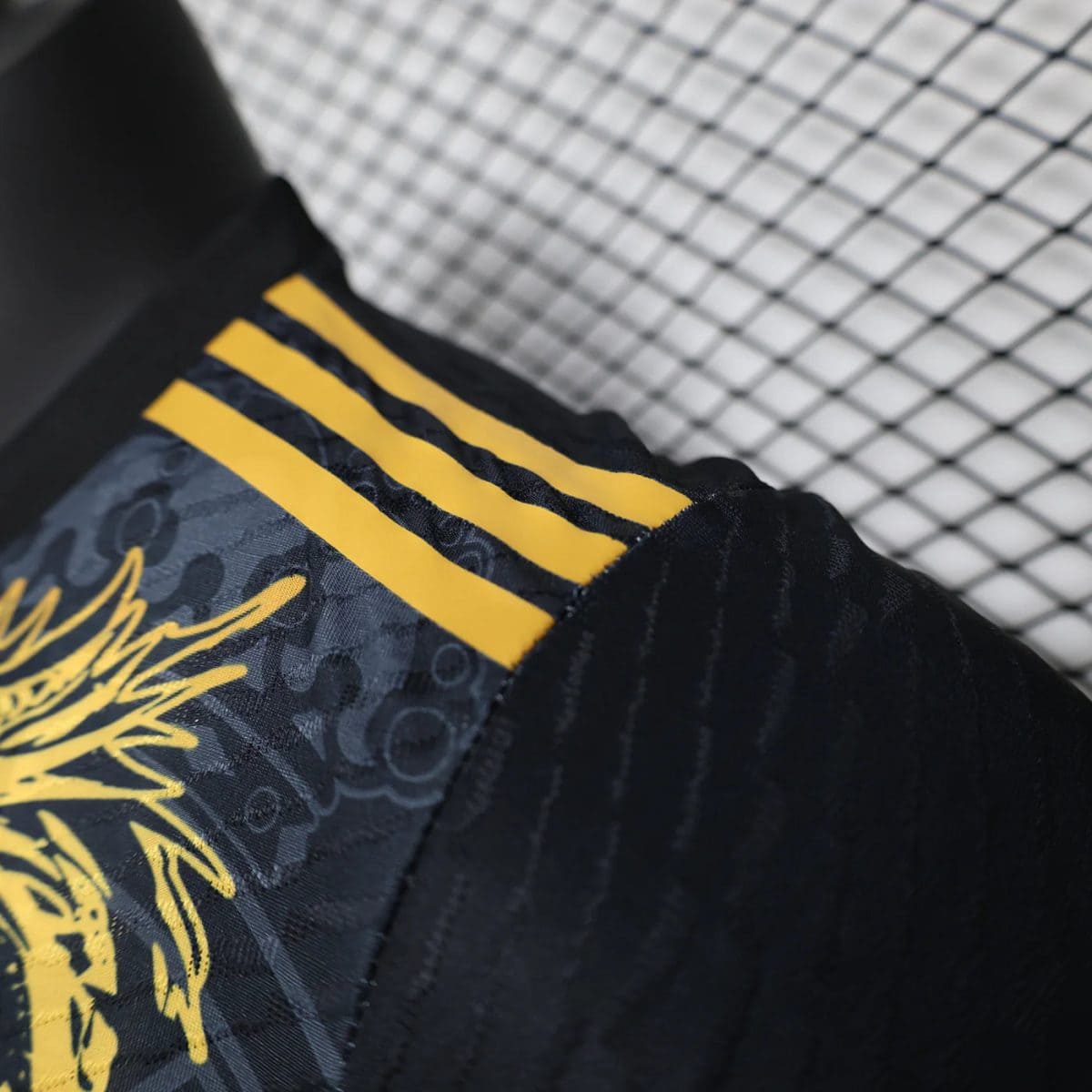 Close-up of dark fabric with three yellow stripes and a gold graphic pattern, likely athletic wear detail, against a chain-link fence background.