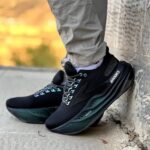 Close-up of black Brooks running shoes with teal laces, worn with light tan pants, foot resting against a textured wall outdoors.