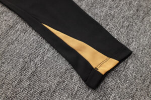 Black pant sleeve with a gold inner lining visible at the cuff on a gray carpet backdrop, highlighting the contrast seam design.