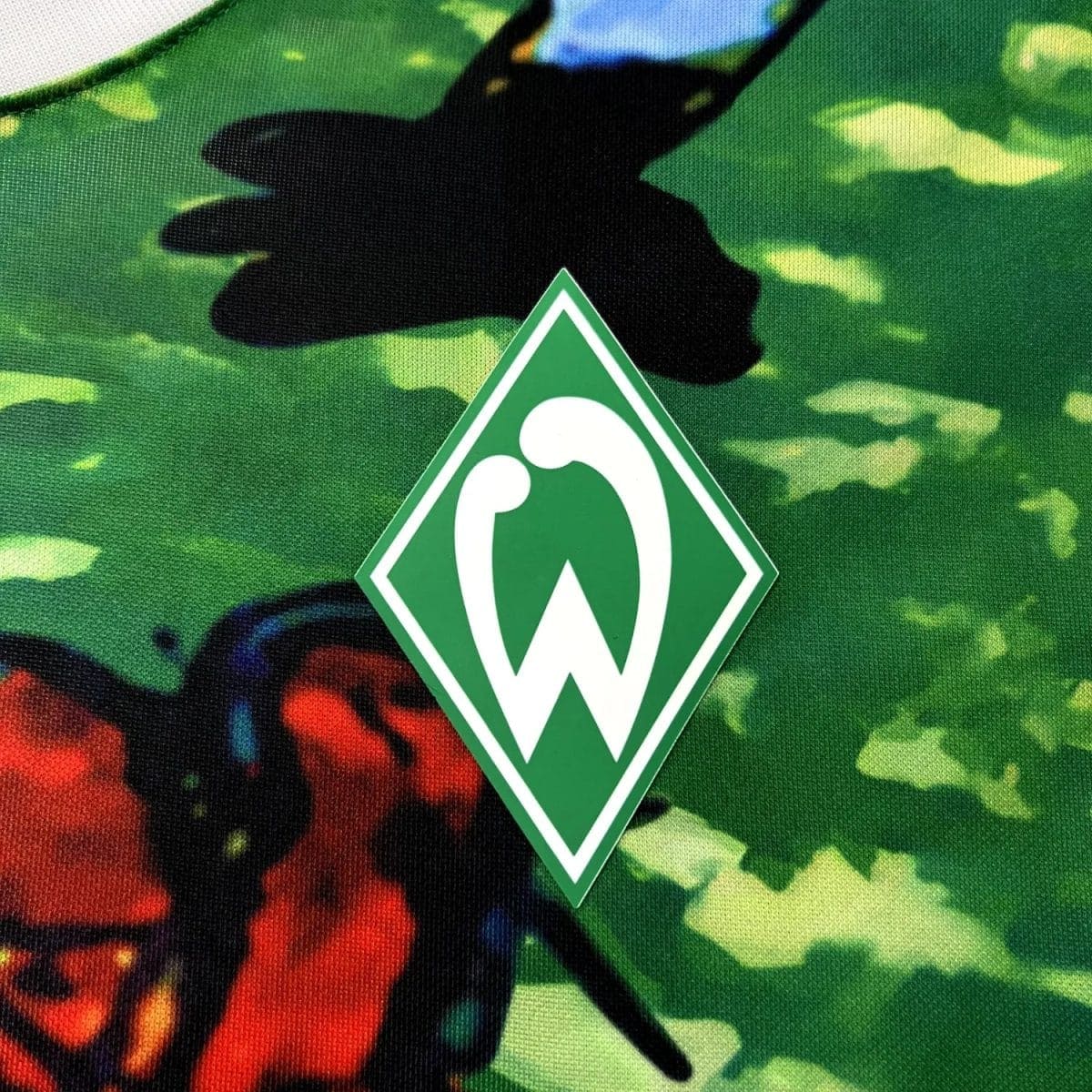Werder Bremen club logo on a green camouflage fabric, close-up of the diamond-shaped green crest with a white 'W'.