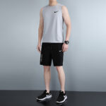Man standing in a light gray sleeveless Nike top, black shorts, and black sneakers with white soles.