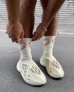 Person kneeling on a street, wearing white sculpted slip-on shoes and white socks with red lettering, adjusting the socks while wearing rings and bracelets.
