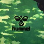 Close-up of a black hummel bee logo with the word 'hummel' on green camouflage fabric.