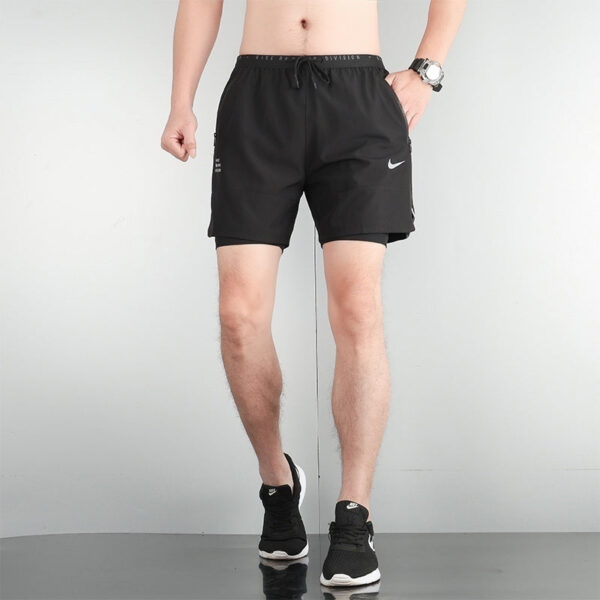 Male model wearing black Nike athletic shorts and black running shoes, standing against a light gray wall.