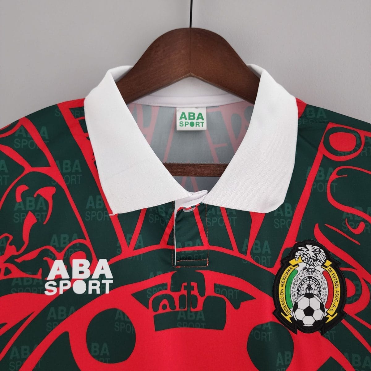 Close-up of a green and red ABA SPORT polo on a wooden hanger with a white collar and a badge showing a soccer ball emblem at the bottom edge.