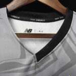 Close-up of a gray New Balance athletic jersey on a wooden hanger, showing the V-neck collar with black trim and NB logo inside.