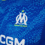 Close-up of a blue Olympique de Marseille jersey with the club crest (interlaced OM and yellow star) and the slogan 'DROIT AU BUT' beneath.
