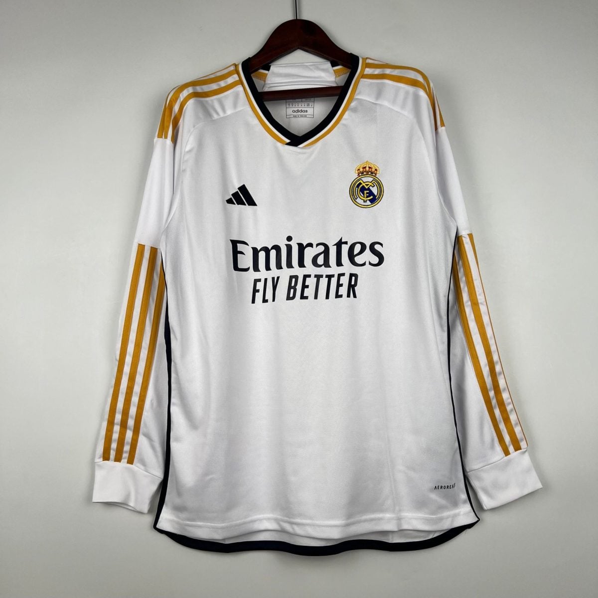White Real Madrid football jersey hanging on a wooden hanger, with gold shoulder stripes and the club crest visible.