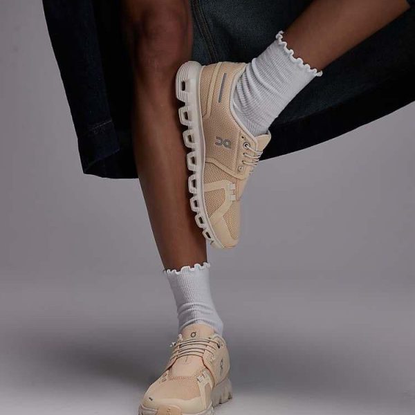 Beige athletic sneakers with chunky white soles, worn with white ruffled socks and dark pants, shown in a posed leg-crossing stance.