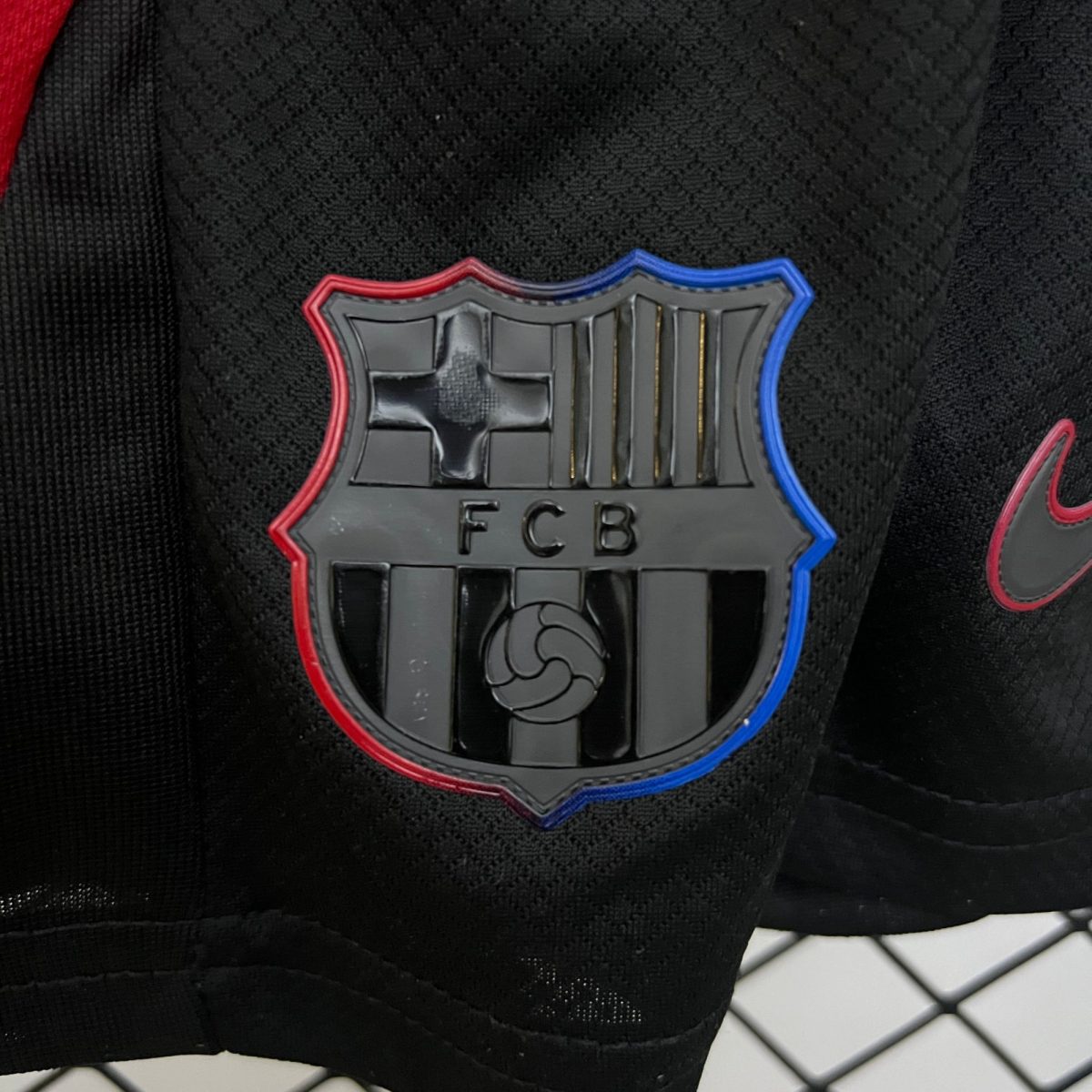 Close-up of a shield-shaped badge with red and blue trim on black fabric, partly showing letters 'FCB'.