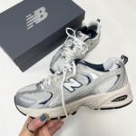 1f16314e-d0b2-4075-8030-51a3cbf07b3d | UAE SPORT STORE Silver and white New Balance 530 sneakers with blue accents, placed next to a gray NB box.