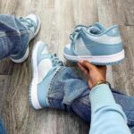 Person wearing light blue Nike sneakers sits on a wooden floor, holding one shoe in hand. You can see denim jeans and a bracelet on the wrist.