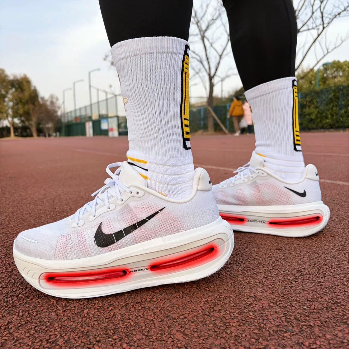 1f7f9c1f-75b2-47b1-9d5f-a1825aeb28af Pair of white Nike running shoes with visible red air-sole units, worn with tall white socks on a red running track.