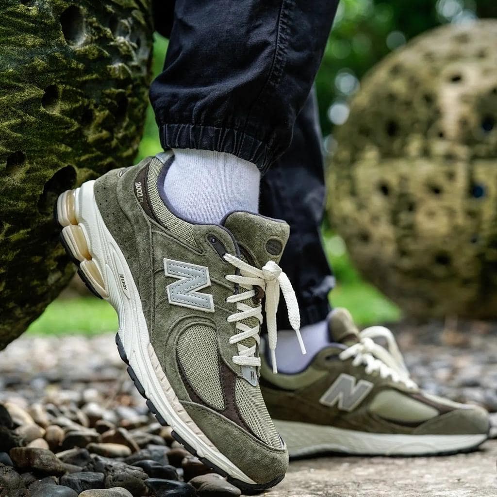 1fa7593a-bc11-4eda-91be-ccda1e057c66_e566efd9-99fd-4286-82c5-b77057b56dd8 | UAE SPORT STORE Olive green New Balance sneakers with white laces, foot resting on rocky path outdoors.