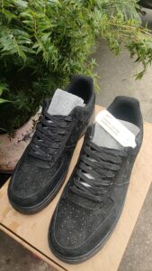 Pair of black sneakers with gray tongues and laces resting on a wooden surface beside green foliage.