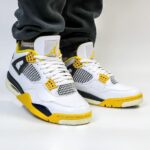 White Air Jordan-style sneakers with yellow accents and black detailing on a white surface, worn by someone standing in gray pants.