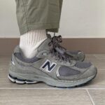 Grey New Balance athletic sneaker with white sock and olive pants, shown from the side on a tiled floor, 2002R model detail visible