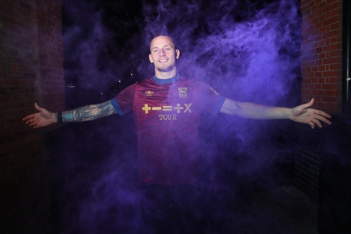 Man in a maroon sports jersey stands with arms outstretched through purple smoke in a brick alley at night.