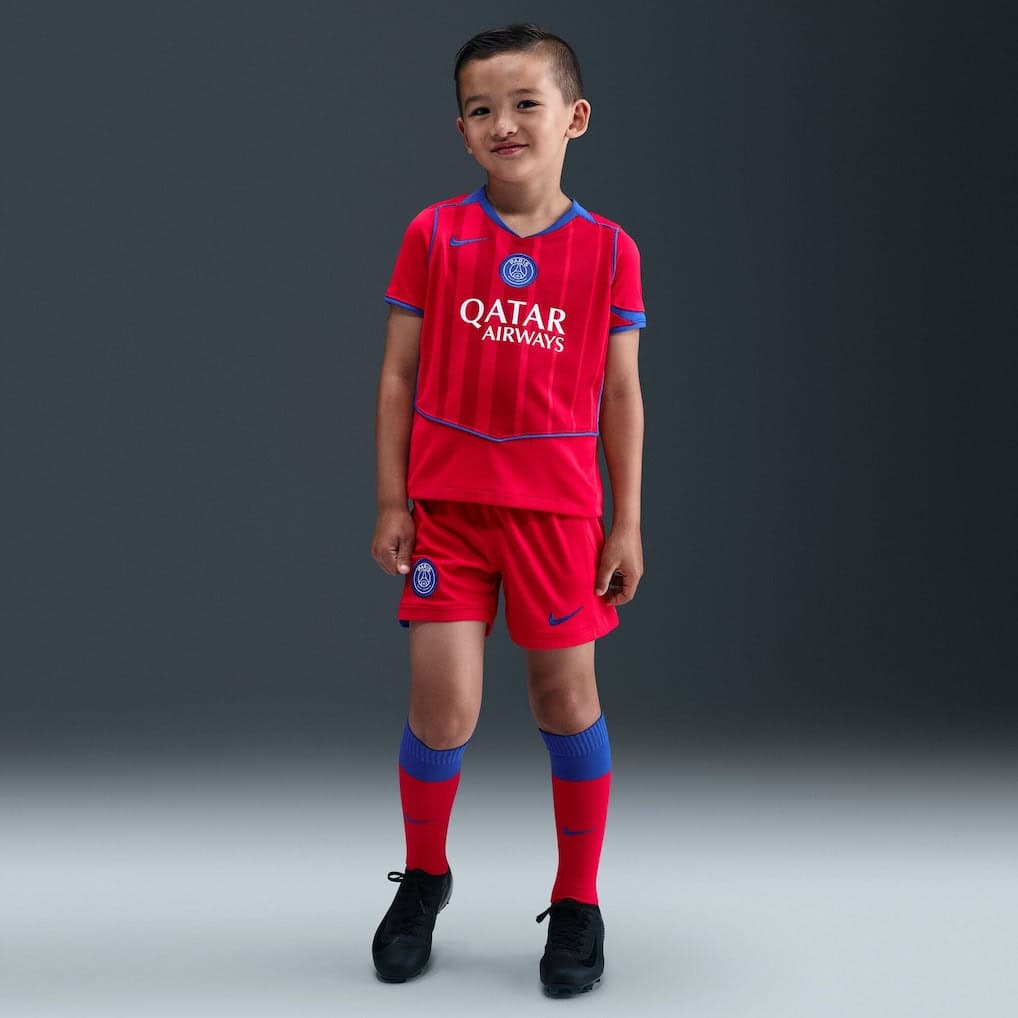 1q | UAE SPORT STORE Young boy in a red PSG-style soccer kit with blue trim, sponsor logo 'Qatar Airways', standing against a gray backdrop.