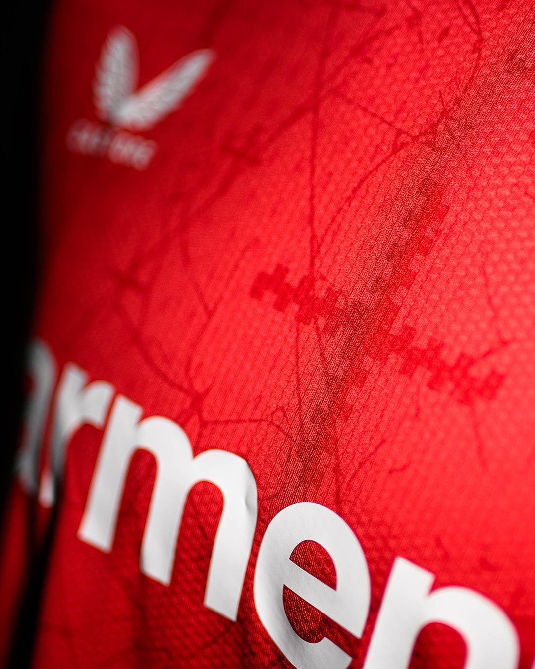1wZC76f2YBjLiGX | UAE SPORT STORE Close-up of a red jersey fabric with a white logo and large white lettering, featuring scribbled signatures or abstract patterns.