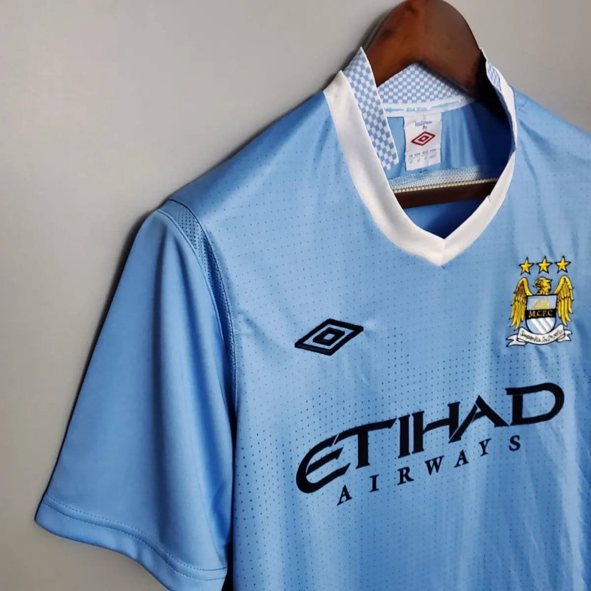 2012-Manchester-City-Retro-Home-Short-Sleeve-Jersey5 | UAE SPORT STORE Light blue Manchester City football jersey on a wooden hanger, white collar and sponsor text Etihad Airways.