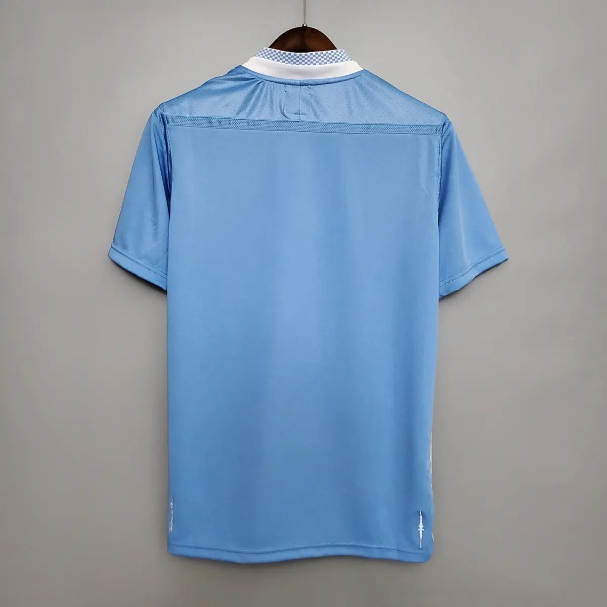 2012-Manchester-City-Retro-Home-Short-Sleeve-Jersey8 | UAE SPORT STORE Light blue short-sleeve polo shirt on a hanger, back view with a white and checkered collar edge visible at the top of the neck area.