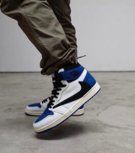 Close-up of a person mid-step wearing white, blue, and black high-top sneakers (Air Jordan 1) with brown pants in an indoor setting.
