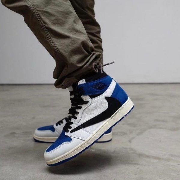 Close-up of a person mid-step wearing white, blue, and black high-top sneakers (Air Jordan 1) with brown pants in an indoor setting.