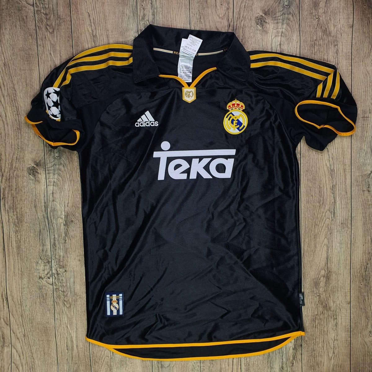 Black Real Madrid football jersey with gold piping and stripes on the shoulders, featuring the club crest and Adidas logo on a wooden floor backdrop