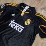 Black Real Madrid football jersey with yellow piping and crest lying on a wooden floor, Adidas logo visible, with care tags attached.