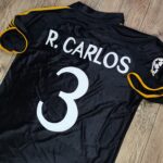 Black sports jersey with orange piping, showing a large white number 3 and the partial name 'R. CARLOS' on the back.