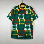 Green sports jersey with a bright yellow collar featuring a bold geometric pattern in red, yellow, white, and dark green; circular club badge on the chest.