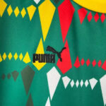Close-up of green fabric with geometric diamond shapes in yellow, red, and white, featuring the black PUMA logo (wordmark with jumping cat).